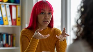 Woman looks like she is explaining something to another woman. Only part of the shoulders and hair of the other woman can be seen and they are blurred. Shelves with books and folders are shown in the background.