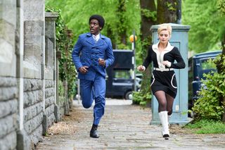 The Doctor (Ncuti Gatwa) and Ruby (Millie Gibson) run down the street towards the camera in 60s attire - he's wearing a navy blue double-breasted pinstripe suit, she's wearing a mod-style black-and-white dress