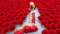 A person in a conical hat arranges bright red incense sticks laid out in circular patterns on the ground