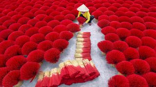 A person in a conical hat arranges bright red incense sticks laid out in circular patterns on the ground