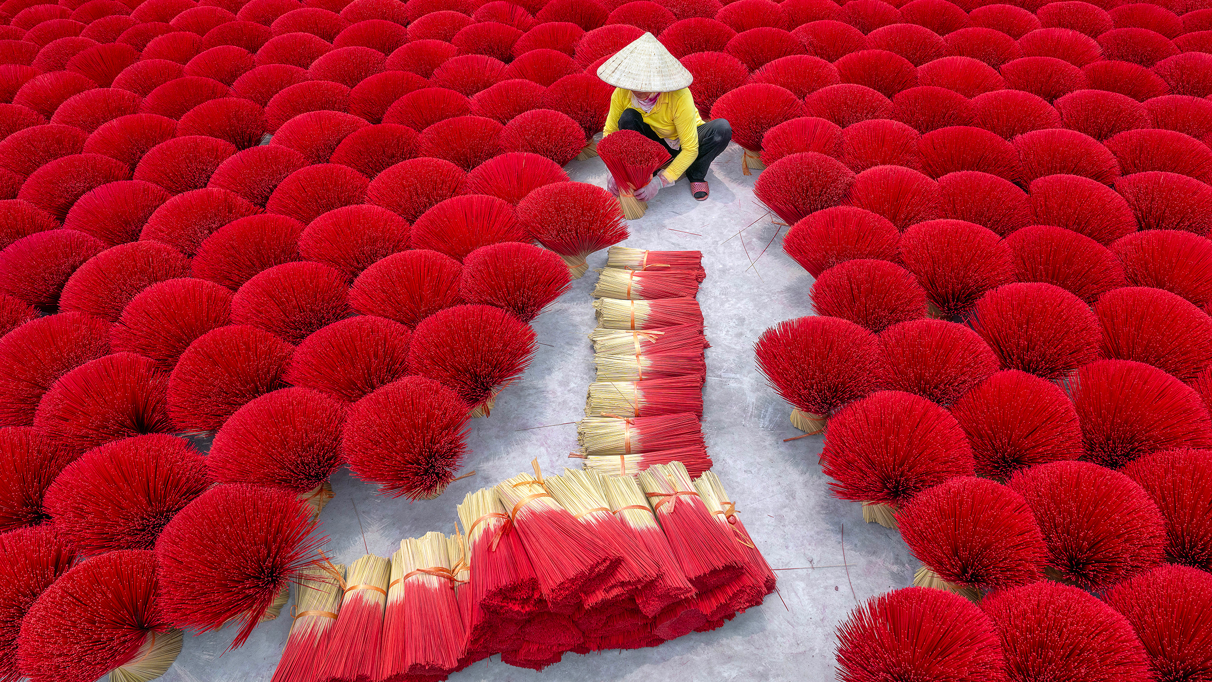 A person in a conical hat arranges bright red incense sticks laid out in circular patterns on the ground