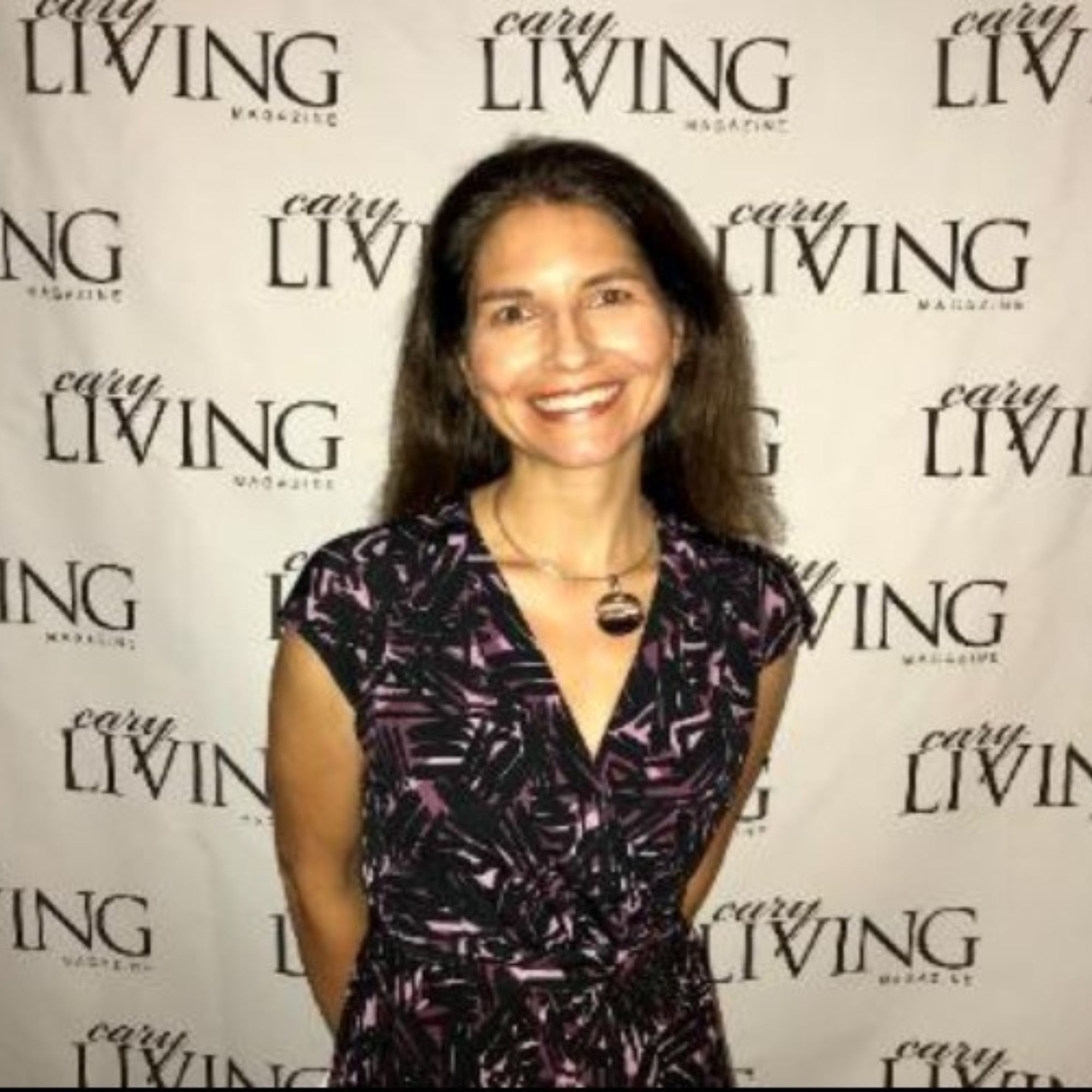 A picture of Amy Bell, a woman with brown hair wearing v-neck navy blue dress standing in front of a white background with black logos saying 'Cary Living' on them