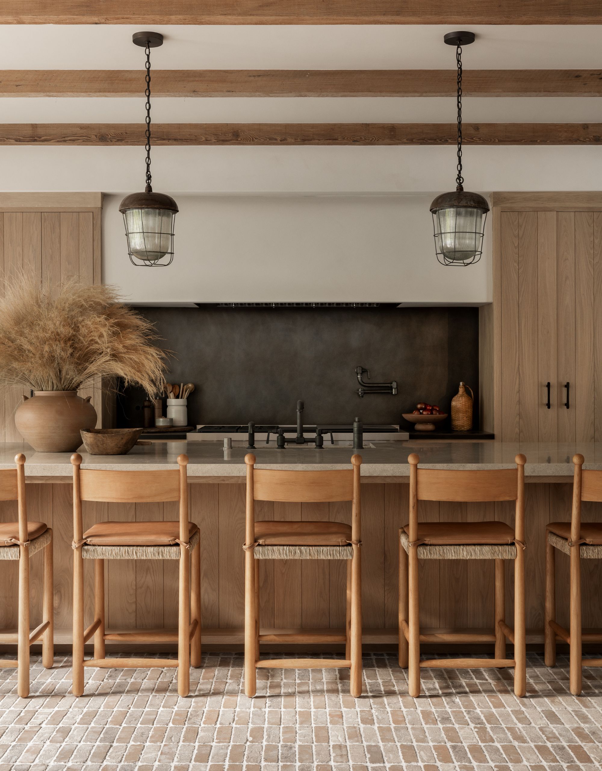 A modern kitchen with wooden cabinetry, a brick flooring, a black textured backsplash in front of the large island with wooden stools with two pendant lights above it and a large vase of foliage