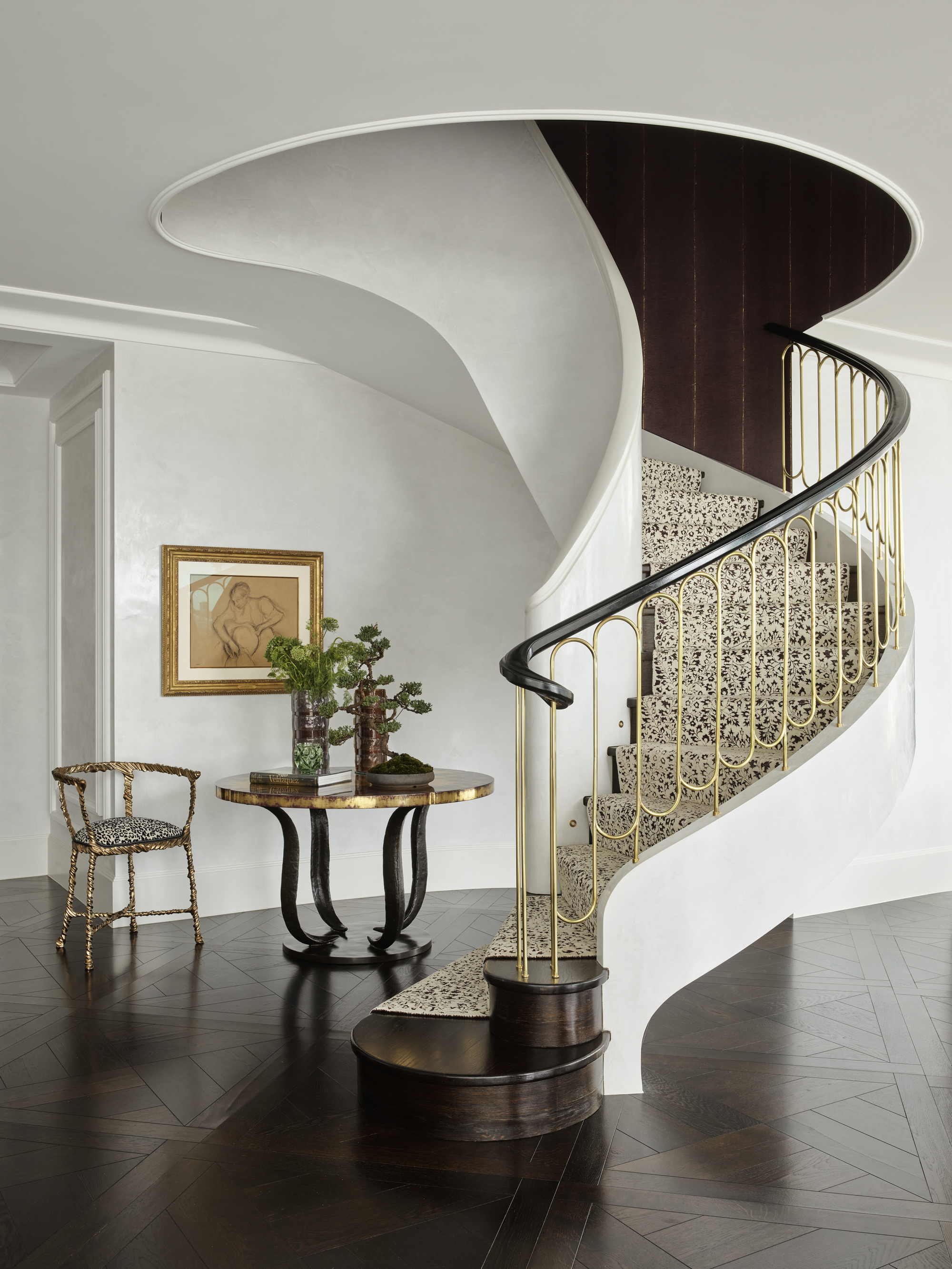 traditional entryway with curved staircase with silk runner and gold balustrade, dark parquetry flooring, with a chair and round table styled with a vase of flowers