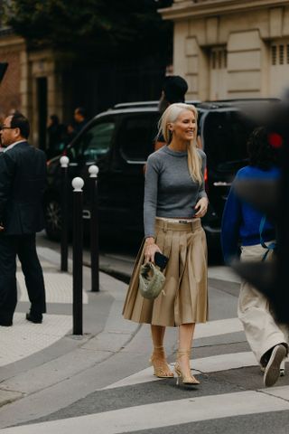 A woman at Paris Fashion Week wearing a gray pullover sweater, tan pleated midi skirt, and gold heels