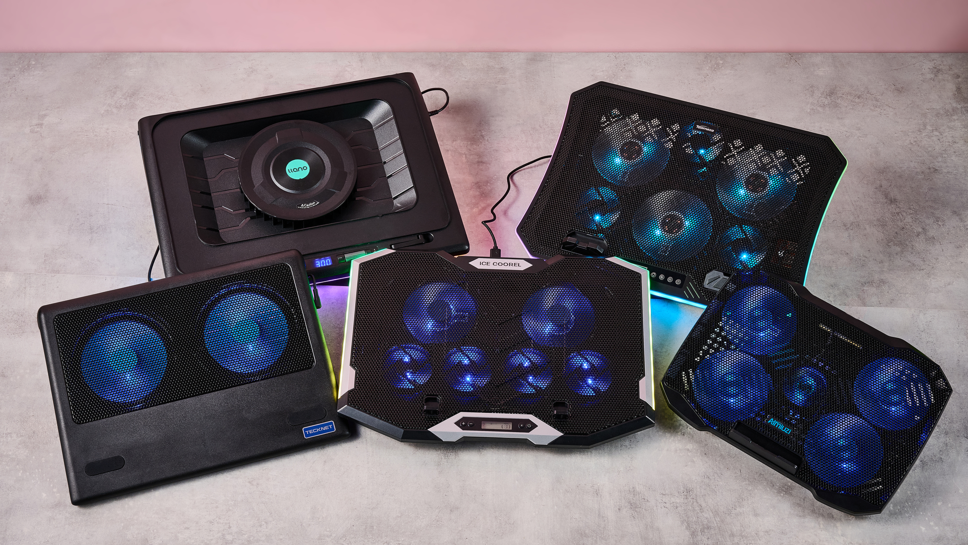 Clockwise: the Llano RGB Laptop Cooling Pad, TopMate C12, Aimuzi F2 Plus, ICE COOREL RGB Laptop Cooling Pad and Tecknet N8 on a gray marbled desktop in front of a pink background.
