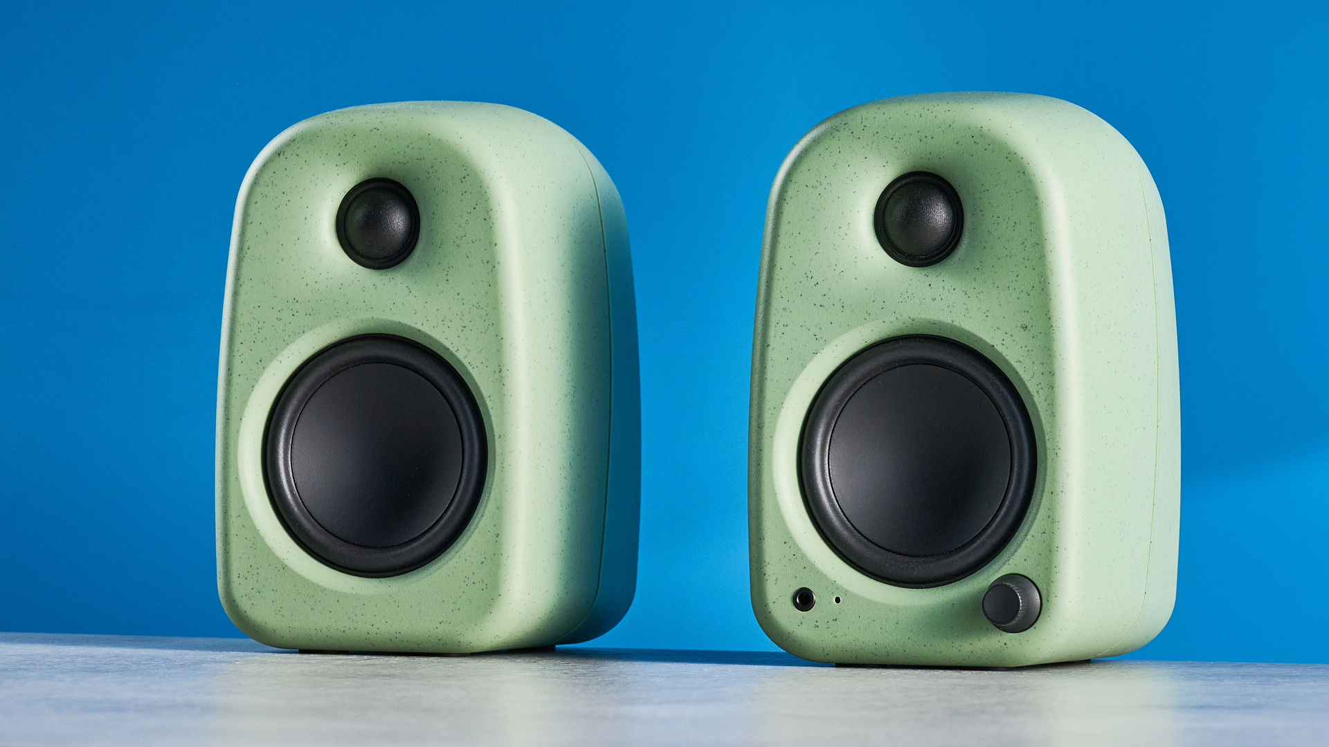 Kanto UKI bookshelf speakers in sage photographed in front of a blue background