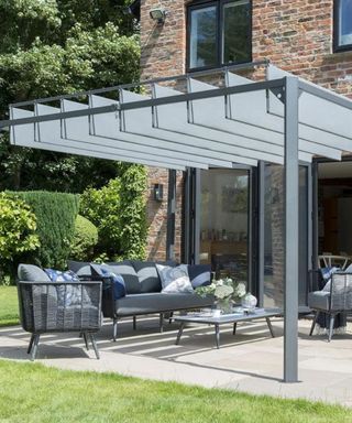 Architectural gray pergola above patio area, with gray garden furniture lounge set-up, on a paved patio next to grass