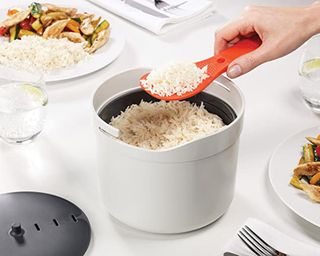 A person serving white rice on table from Joseph Joseph 2 liter capacity microwave rice cooker with red paddle spoon