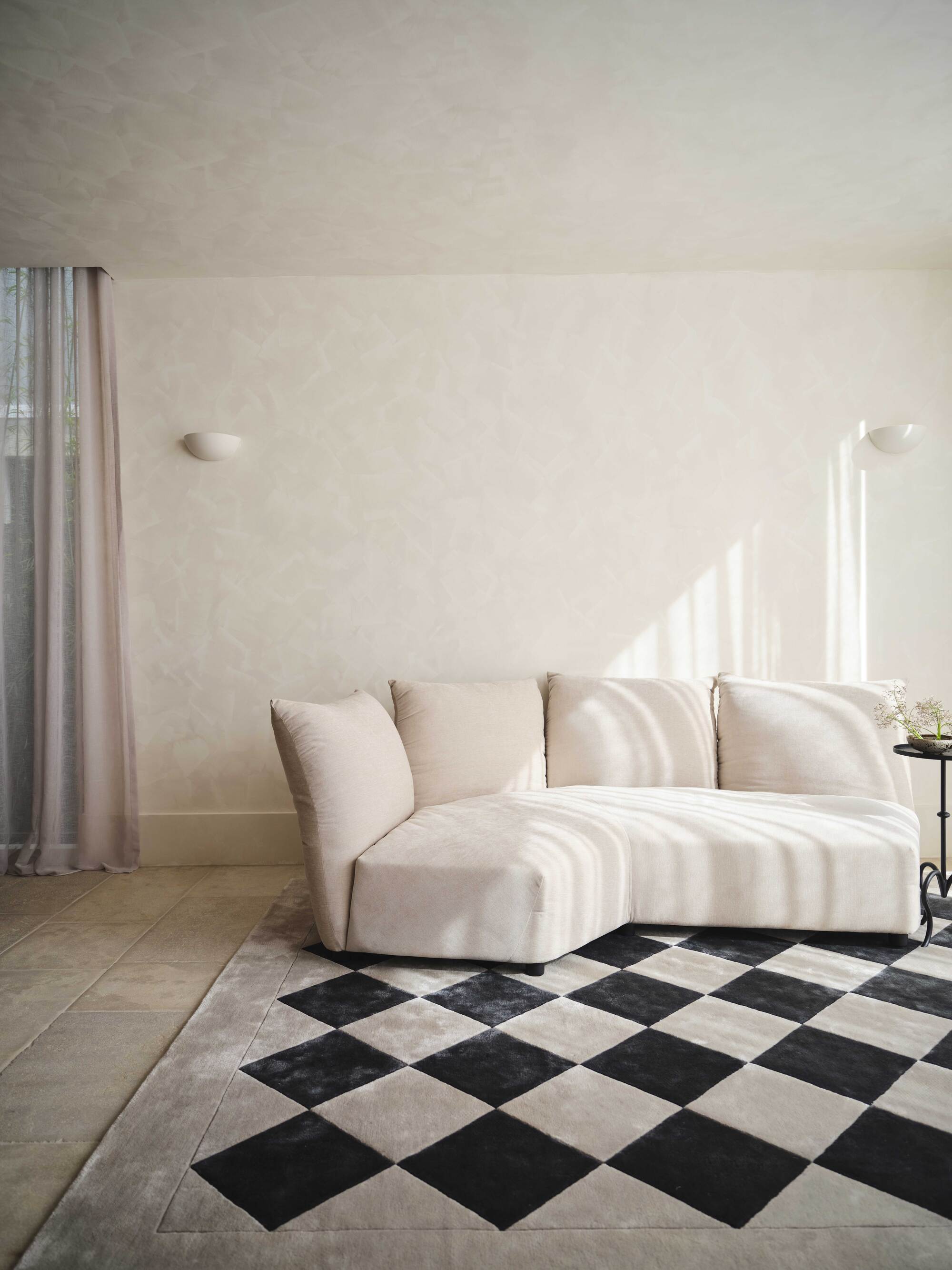 Image of an off-white, limewashed living room. There is a cream, sectional sofa with a black and white checkerboard rug.