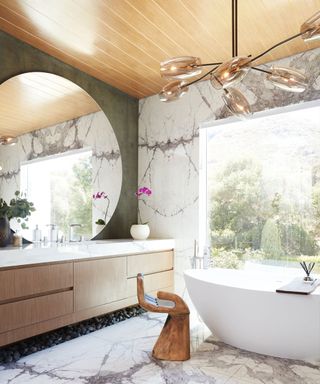 Spa inspired bathroom with wooden paneled ceiling. marble walls, large rounded mirror above vanity area, marble flooring, large white bath beside floor to ceiling window, hand sculpture seat, metal and glass pendant