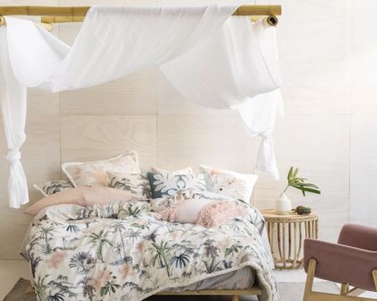 A white and palm print duvet on a bed with bamboo plinths attached to walls and white veil material draped over