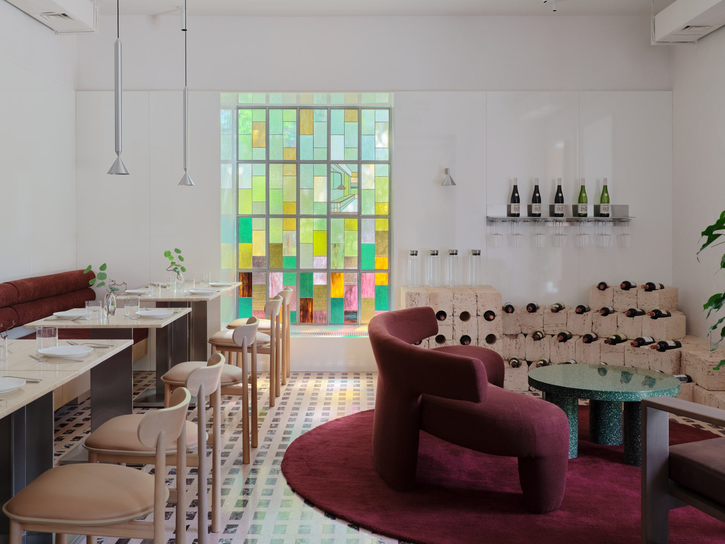 An eccentric dining room with checkered brown and beige floors, pale wooden floors, chrome and wood tables, stained glass windows, rounded velvet red rugs, chunky armchairs, and wine racks.