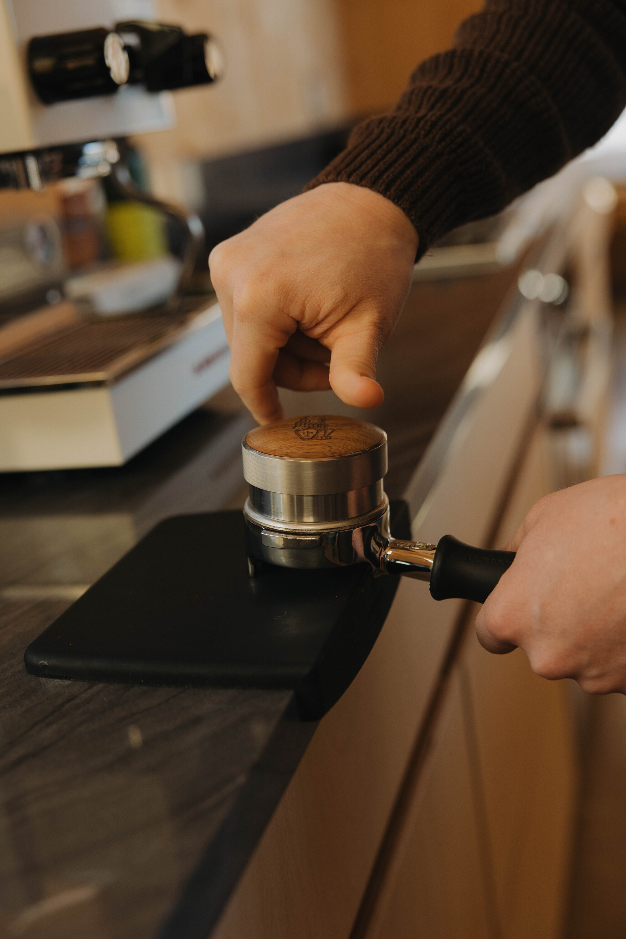 Image of a hand tamping and weighing coffee grounds in a portafilter on a tamping mat on a dark kitchen countertop.