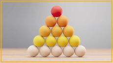 A pyramid of tan, yellow, orange and red wooden balls are stacked on a wooden surface with a blurry gray background and yellow border around the image