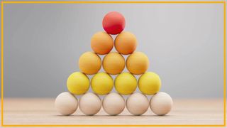 A pyramid of tan, yellow, orange and red wooden balls are stacked on a wooden surface with a blurry gray background and yellow border around the image