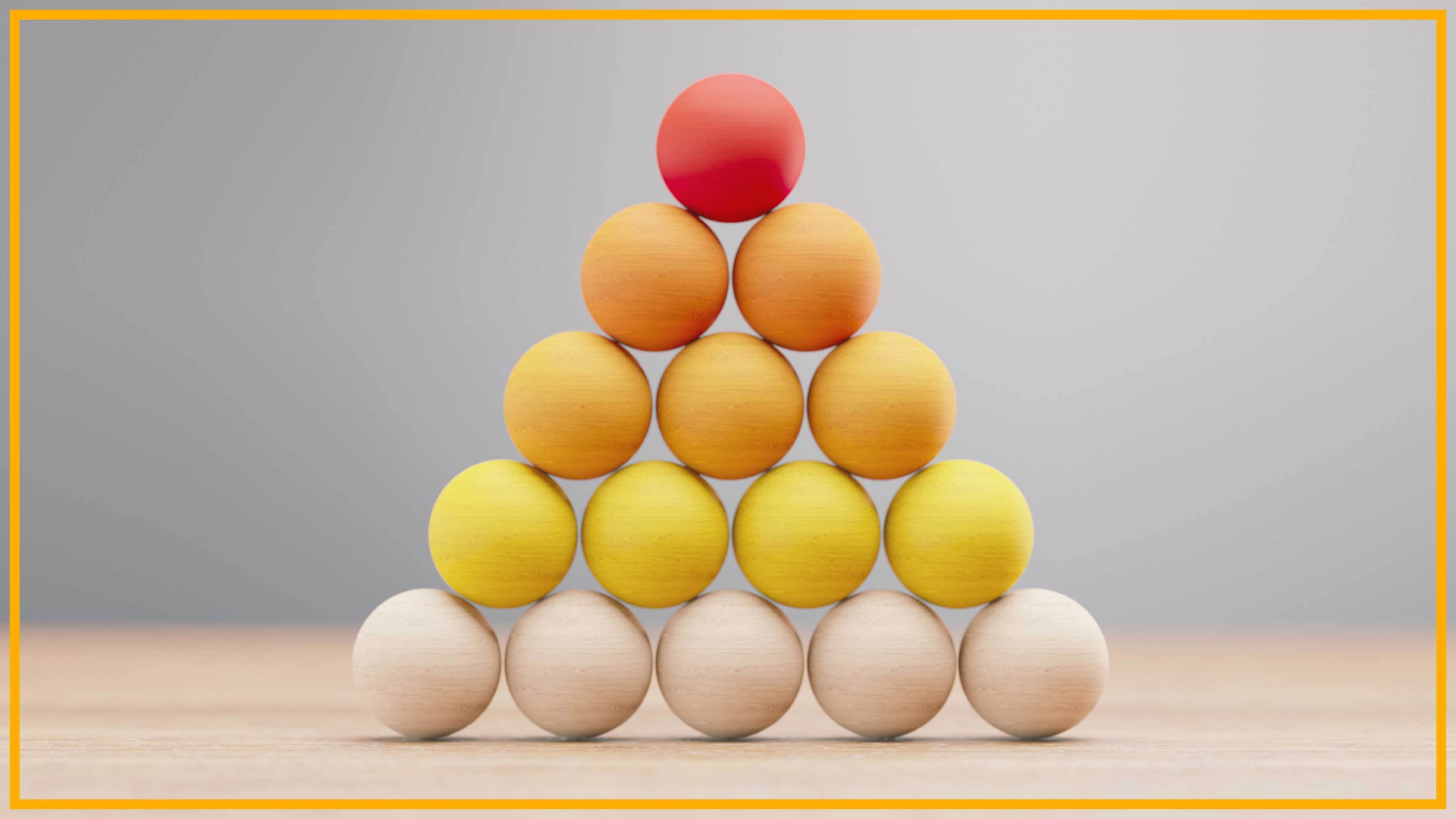 A pyramid of tan, yellow, orange and red wooden balls are stacked on a wooden surface with a blurry gray background and yellow border around the image