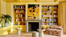 yellow living room with built-in shelves, checkered fireplace, large artwork above it, a yellow console with potted plant, three side tables, a patterned sofa and timber floorboards