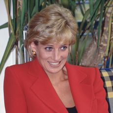 Princess Diana has short blonde coiffed hair and wears a bright red jacket with a black top underneath and looks sheepish