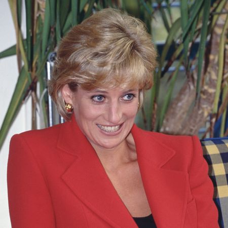 Princess Diana has short blonde coiffed hair and wears a bright red jacket with a black top underneath and looks sheepish