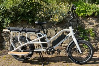 The Mycle Cargo Electric bike is one of the best options on a budget. This one is show side on in front of a stone wall with greenery