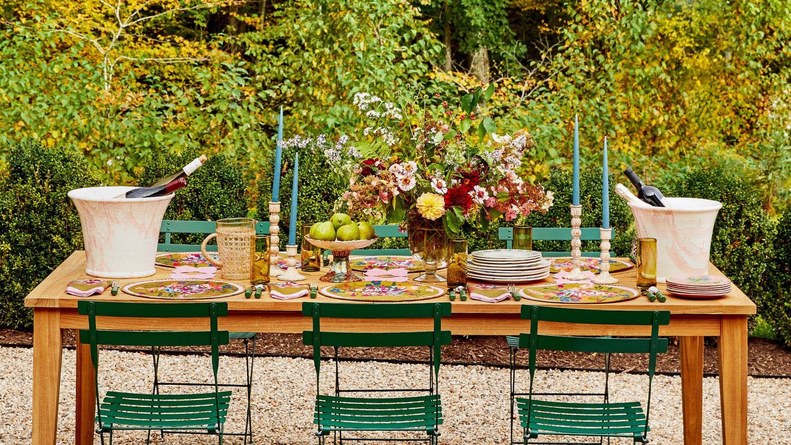 Summer tablescape with blue candles, large floral arrangement, colourful placemats and napkins, white ice buckets all on top of a wooden dining table with green iron chairs 