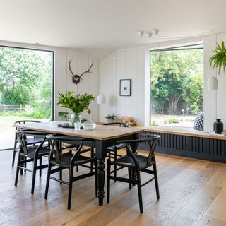 Open plan dining room with wood dining table with painted black legs, black wishbone chairs, wooden flooring, white panelled walls, antler wall art