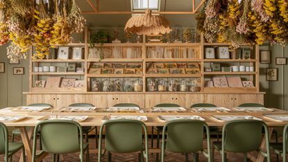 Room with sage green walls, long wood dining table and wood bookcase with rattan pendant light, dried hanging flowers and green chairs