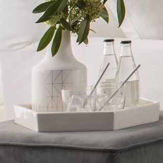 Shiny white hexagonal trinket dish with water glasses, two bottles of water and a white and gold geometric patterned vase with green leaves and budded plant