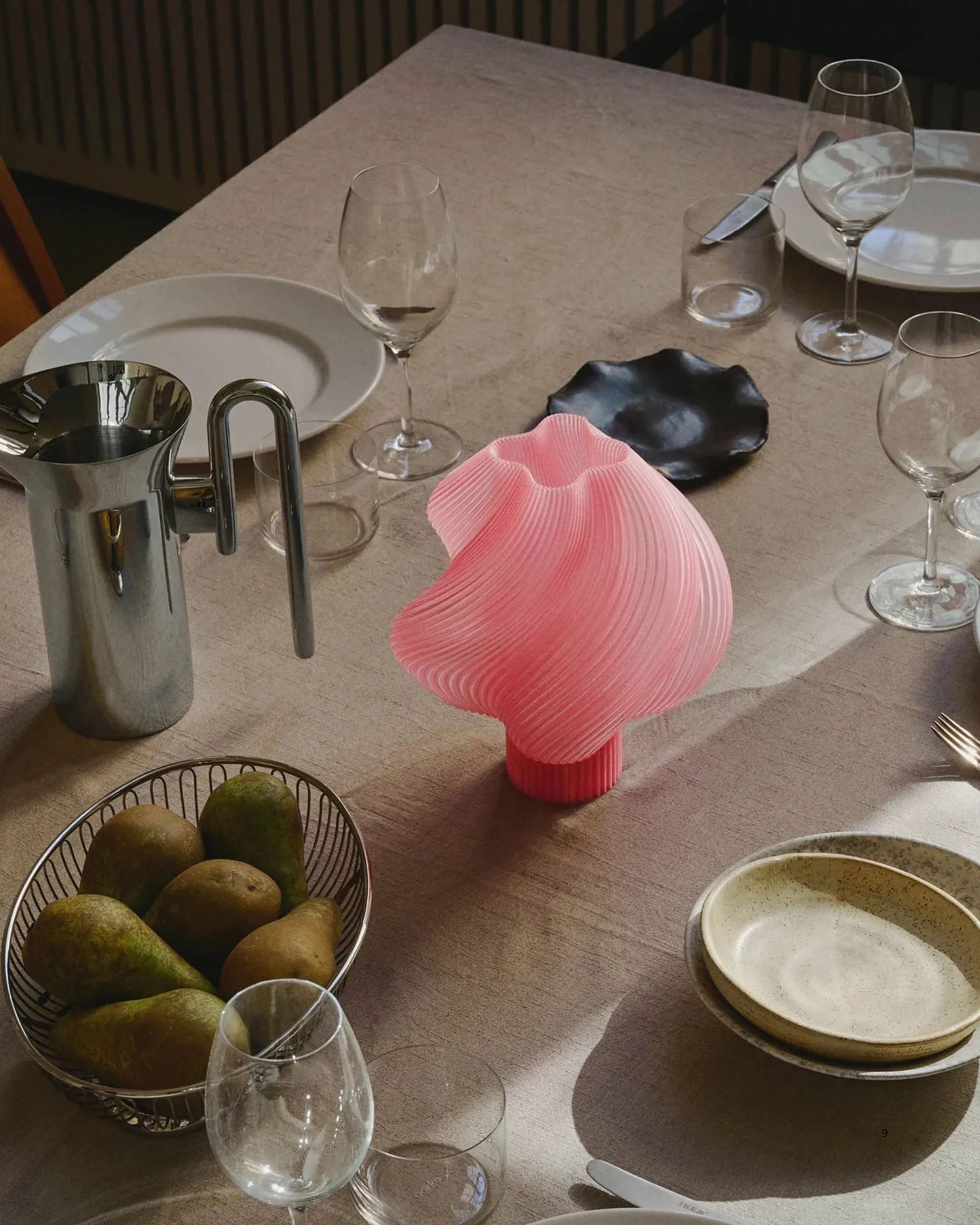 A close-up image of a table set with a beige table cloth, wine glasses, a bowl of pears, and a chrome water jug. There is a pink, swirly table lamp in the center of the table.