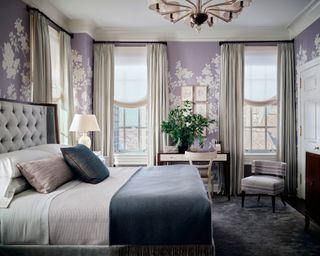 bedroom with lilac floral wallpaper, white drapes, light gray button back winged bed with blue throw and white dressing table and chair