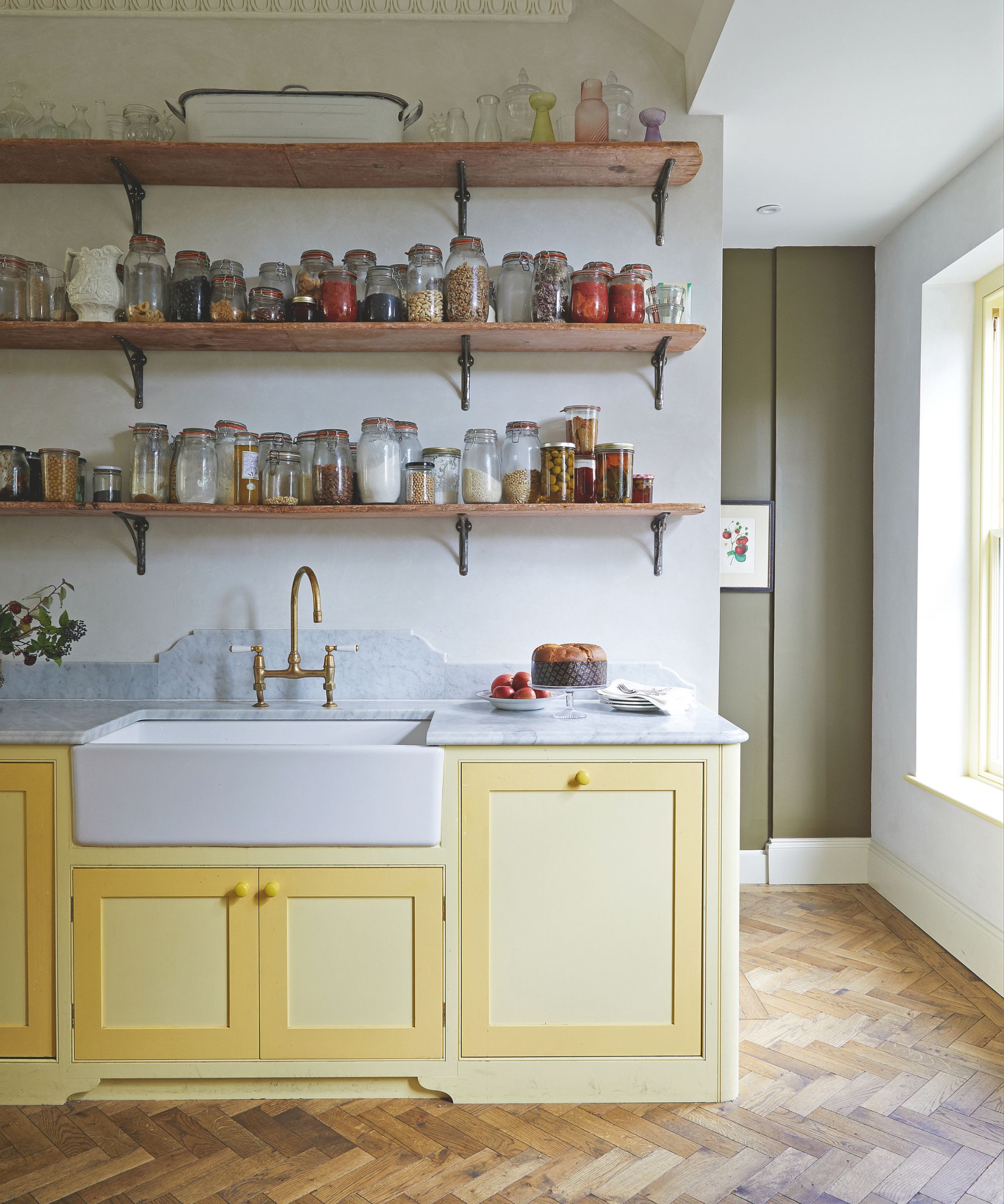 Kitchen with yellow cabinets, wooden herringbone floor and open shelves on the wall behind the sink, showing dozens of mismatched glass storage hars