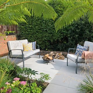 Paved patio area with contemporary garden sofa and armchair next to a small firepit with overhanging palm trees