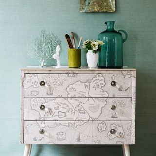 a chest of drawers covered in a map print in front of a green papered wall