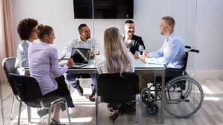 A multi-ethnic, mixed gender team of six colleagues sitting round a meeting room table listening to one member speak, who is in a wheelchair