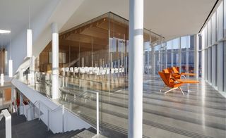 An open space in the building, with a sitting area featuring orange chairs and a table (facing the glass panels looking out). Behind the chairs is a conference room with white chairs. On the left is a peek of the grey stairs going down
