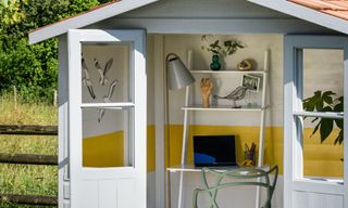 A grey painted garden shed converted into a home office, open to reveal yellow painted interior walls and floor, a ladder desk, green chair and brass floor lamp