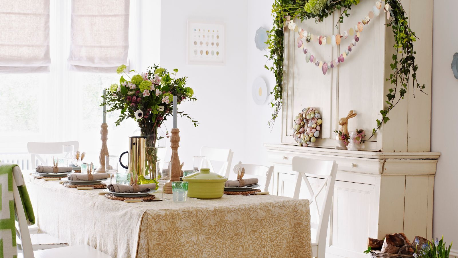 Table set for Easter with pale green and yellow theme, vase of flowers in the middle and a dresser next to the table with an Easter wreath and garland hanging on it