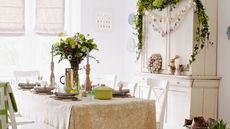 Table set for Easter with pale green and yellow theme, vase of flowers in the middle and a dresser next to the table with an Easter wreath and garland hanging on it
