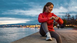 Woman wearing headphones, sitting on a riverbank in workout clothes, checking fitness tracker