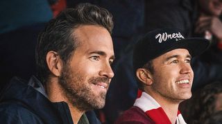 Ryan Reynolds and Rob McElhenney watching a game at the Wrexham stadium in Wales, draped in red and white club colours.