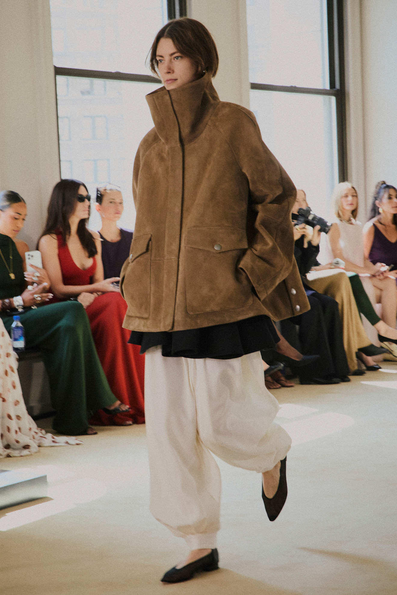 a model at Altuzarra Spring 2026 wearing a brown suede funnel-neck jacket, black tunic, white parachute pants, and black flats