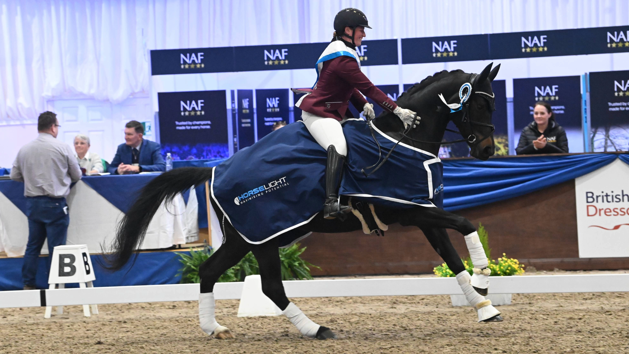 Woman pats a black horse she is riding at a canter. The horse wears a blue winners rug