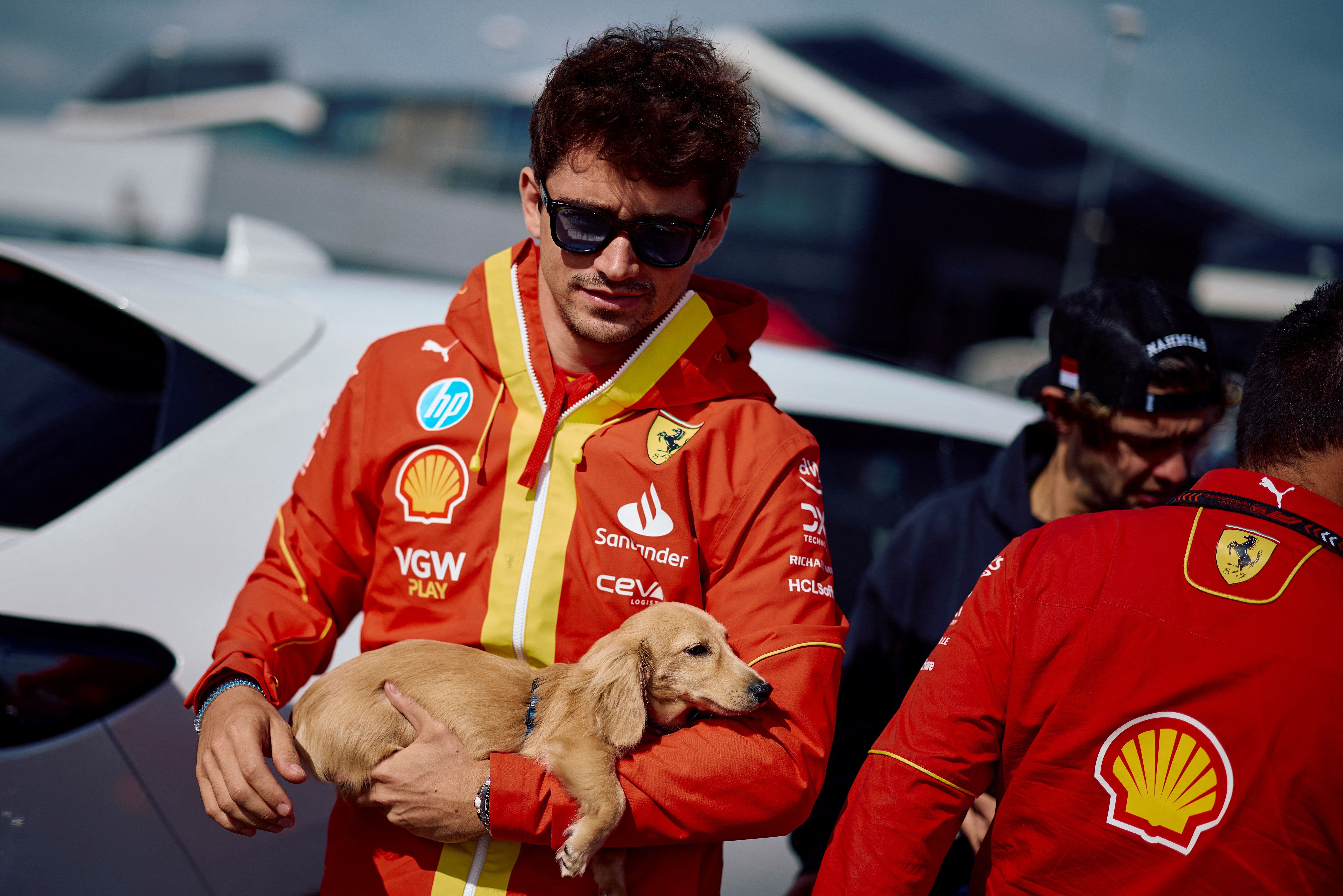 Charles Leclerc in Ferrari uniform wearing sunglasses holding his small golden dachshund puppy. 