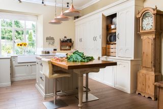 Free standing kitchen furniture in an cream and wood kitchen, and a giant wooden clock to the right, with hanging lights over the island