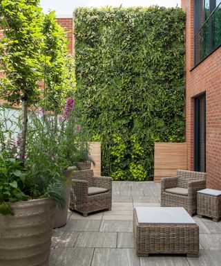 A living wall in a small patio garden beside a tall red brick building, with wicker furniture.