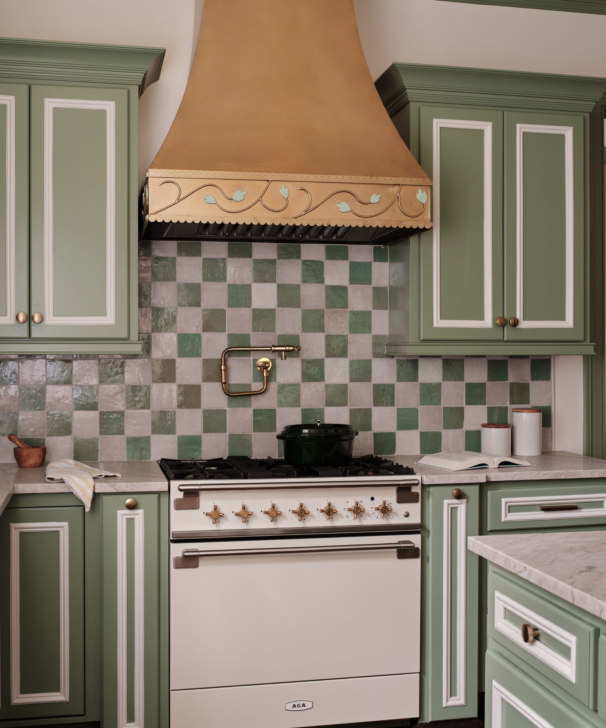 A traditional kitchen with mid-green cabinets with white details, a green and white checkerboard backsplash, a white oven, and a beige terracotta-colored extractor fan.