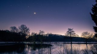 venus shining bright above a lake. trees are silhouetted behind and the sky is a pink purple hue.