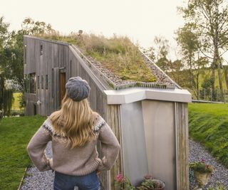 woman with blond hair wearing jumper, jeans and hat stood in front of timber clad modern home with green roof