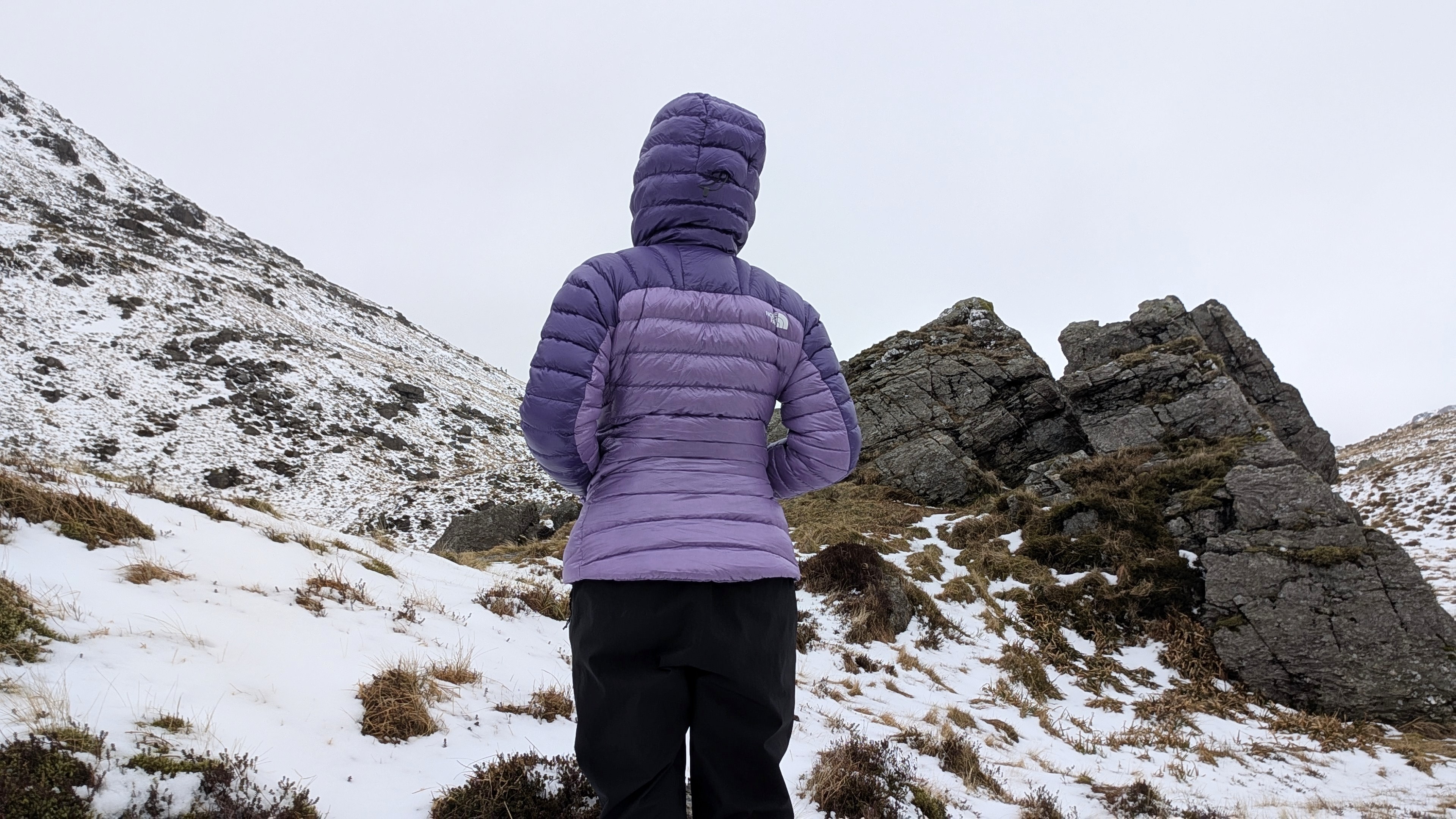 A woman hiker is photographed from behind wearing a purple North Face Summit Breithorn Down Jacket with the hood up